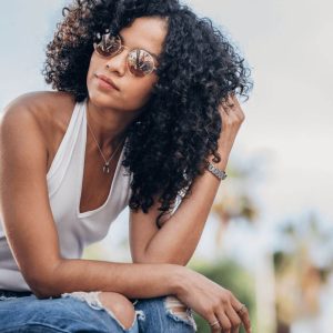 Young woman with dark curly hair, wearing sunglasses, a white tank top and jeans. 